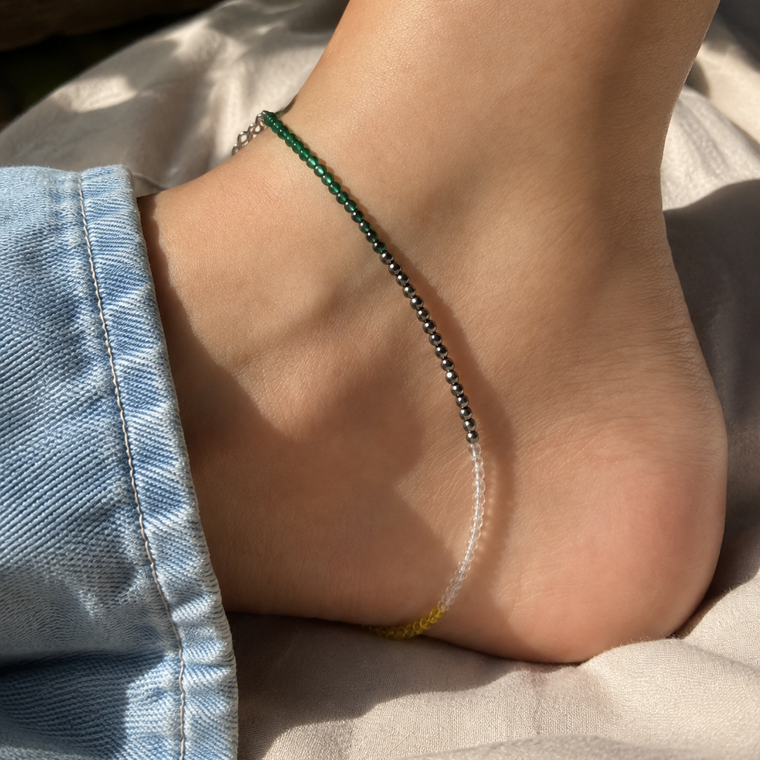 A delicate beaded anklet worn on a person's ankle, featuring green, black, and yellow beads on a silver-toned chain, shown with light blue denim jeans in natural sunlight.