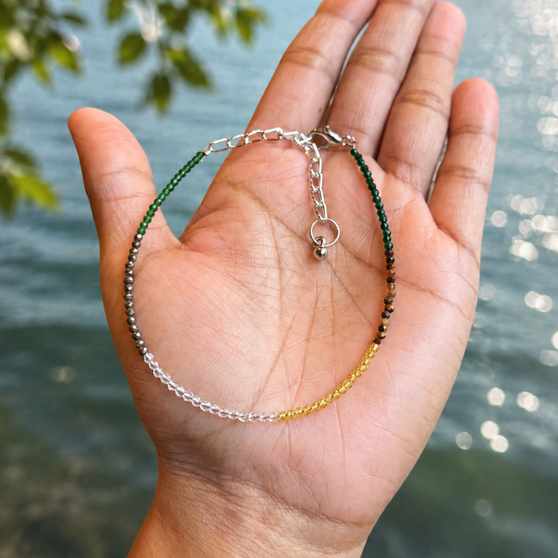 A delicate beaded bracelet with silver, green, and gold beads draped over an open hand, with a blurred lake and forest background.