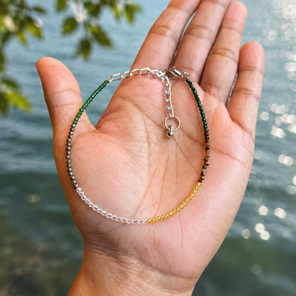 A delicate beaded bracelet with silver, green, and gold beads draped over an open hand, with a blurred lake and forest background.